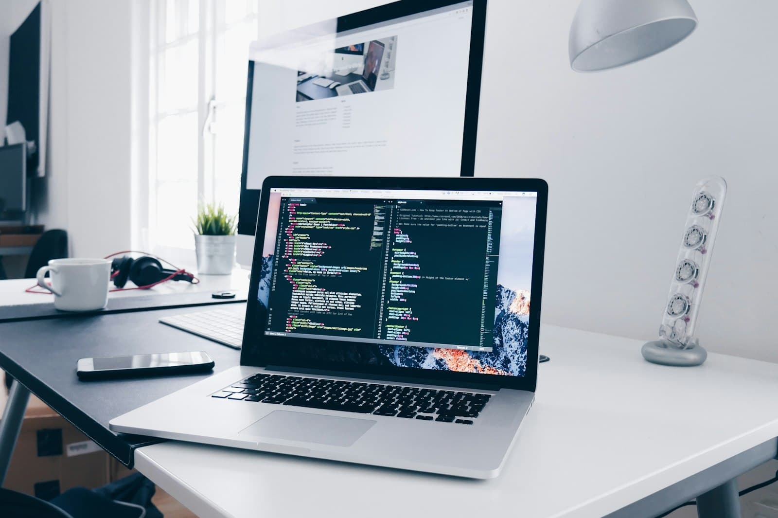 A laptop on a white desk, open to code, with a monitor and a window behind it.