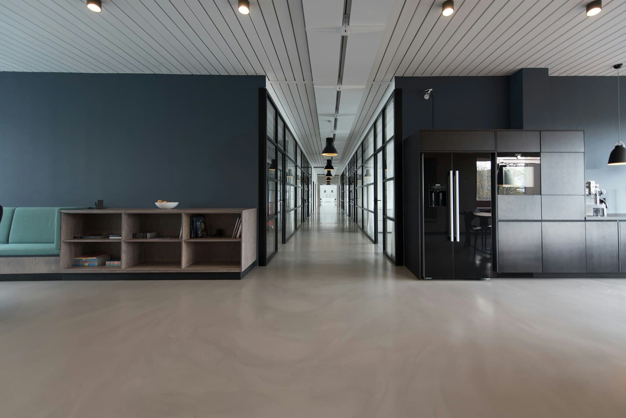 A long office corridor with dark walls, glass rooms, and a pale concrete floor.