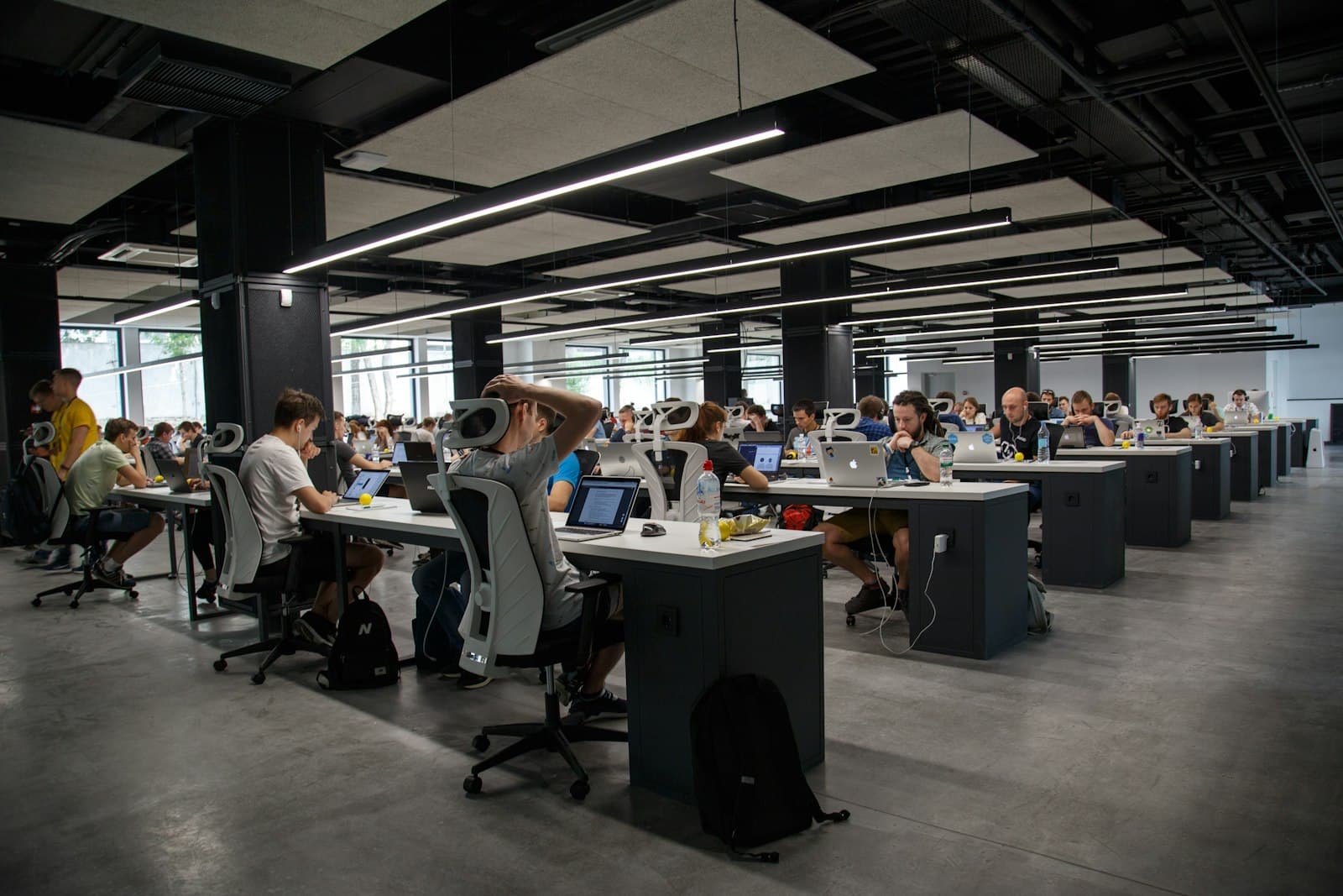 A large open office where people work at laptops under long rows of lights.
